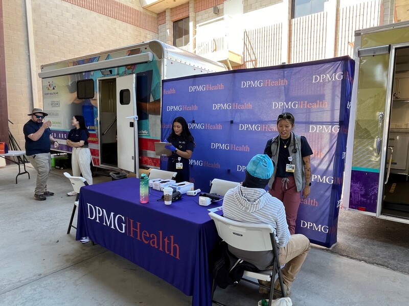 DPMG Health employees stand and talk near the parked mobile units, while some colleagues tend to a farmworker who is seated nearby. Staff Photo