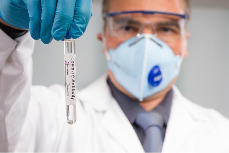 Masked and gloved man in medical gear holds up a vial marked "antibody testing."