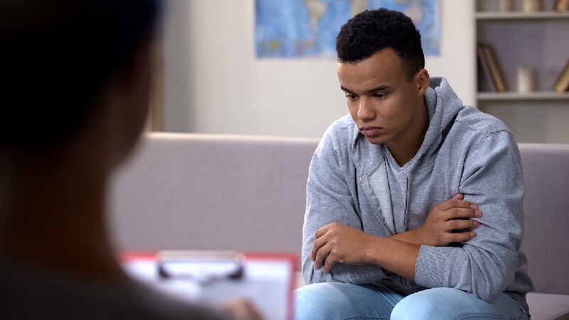 A young black man looks down at the floor during a therapy session.