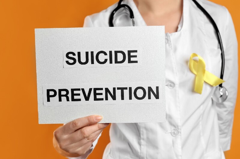 Medical professional in a white coat bearing a yellow ribbon holds a "Suicide Prevention" sign