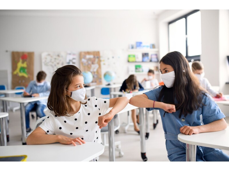 Two masked girls in a classroom greet by touching elbows. Stock Photo