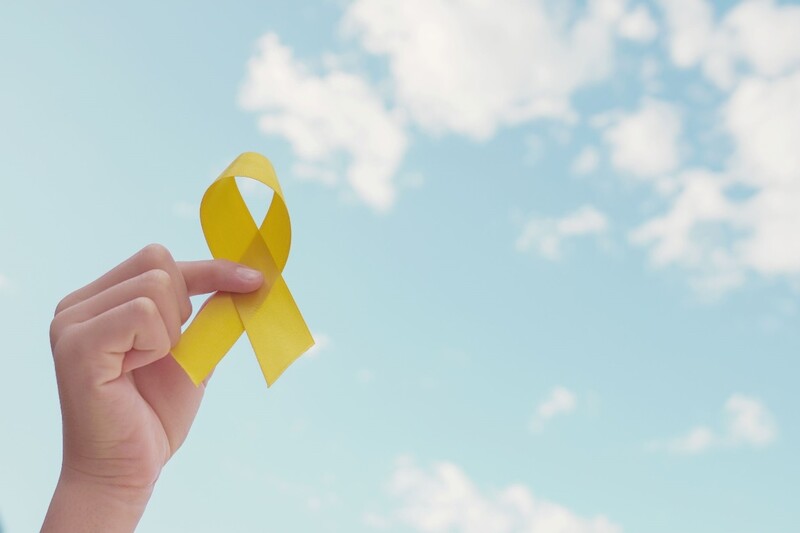 A hand holds up a yellow ribbon with a blue sky with white clouds in the background. Stock Photo