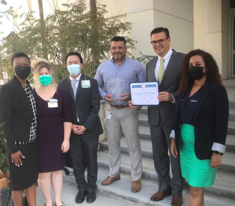 Desert Healthcare District employees hold a plaque and certificate, flanked on both sides by Riverside University Health System employees.