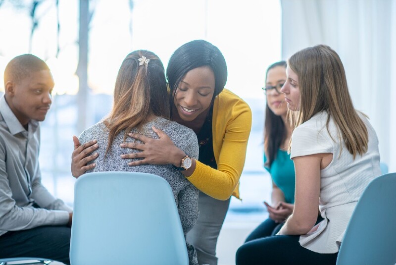 Group support is one resource available to survivors of suicide. Stock Photo A woman hugs another among of group of five people sitting in a circle as a support group. Stock Photo