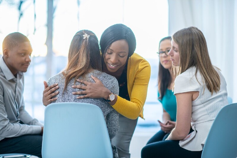 A woman hugs another among of group of five people sitting in a circle as a support group. Stock Photo