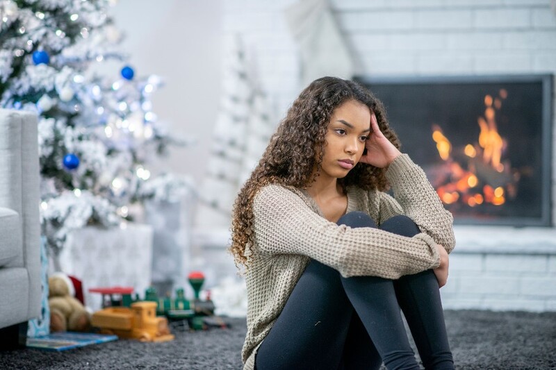 A young woman sits on the living room floor looking despondent, with a Christmas tree in the background. Stock Photo