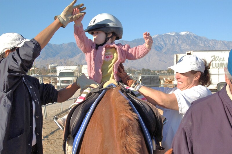 A child wearing a helmet rides a horse with adult escorts walking on both sides of the horse.