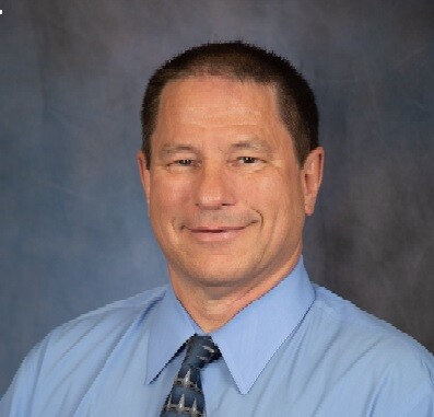 Photo of CAO Chris Christensen smiling and looking at the camera. he wears a medium blue button-down shirt with a darker blue tie. Photo by Lani Garfield