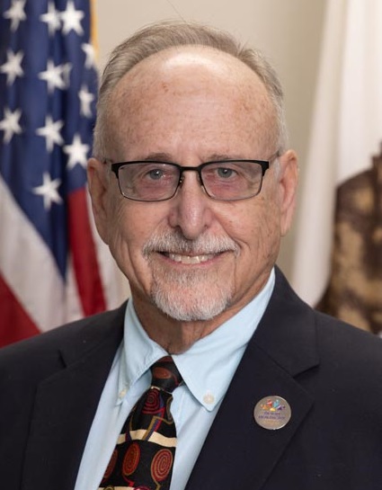 Headshot of Dr. Les Zendle, smiling toward the camera, with the U.S. and California flags behind him. Photo by Lani Garfield