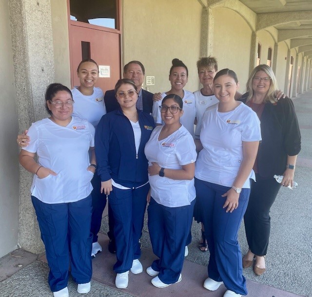 Some of the students participating in the College of the Desert associate degree in nursing program pose with Desert Healthcare District & Foundation CEO Chris Christensen (back row, second from left) and Chief Program Officer Donna Craig (back row, fourth from left) at College of the Desert's Palm Desert campus. Photo by Will Dean Two rows of College of the Desert registered nursing students standing and facing the camera, with agency CEO Chris Christensen, CPO Donna Craig and a female college official standing in the back row with two students.