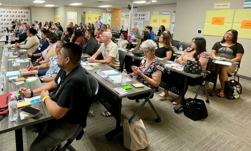A diverse group of people seating at rows of tables facing an unseen speaker.