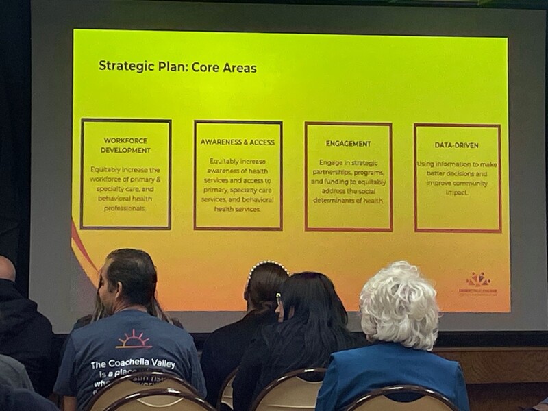 In the foreground, a seated audience from the back. They're looking at (in the background) a bright yellow-green screen that displays the four core areas of the strategic plan. Staff Photo In the foreground, a seated audience from the back. They're looking at (in the background) a bright yellow-green screen that displays the four core areas of the strategic plan. Staff Photo