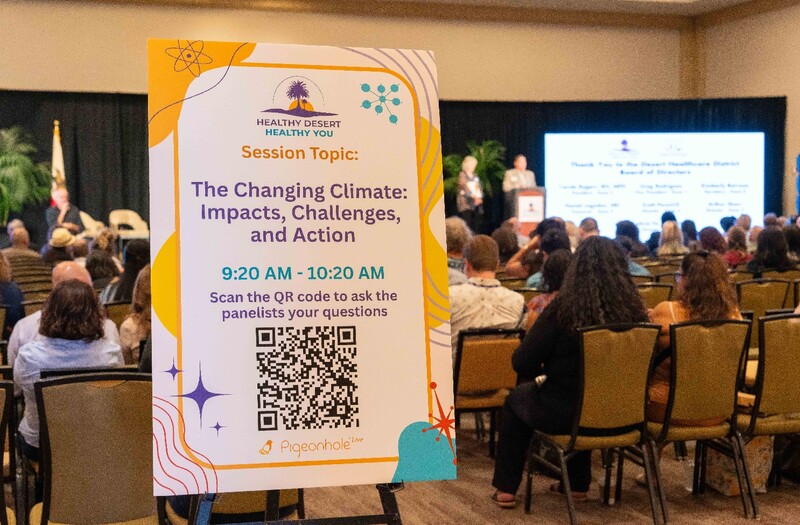 A colorful poster board on an easel announcing a summit session (in the foreground) and the back of a seated audience looking toward the stage in the background. Staff Photo A colorful poster board on an easel announcing a summit session (in the foreground) and the back of a seated audience looking toward the stage in the background. Staff Photo