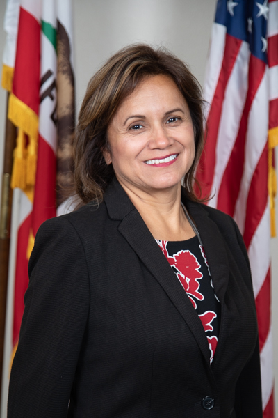 Leticia De Lara, DHCD Board President Board Director Leticia De Lara smiling with U.S. and California state flags in background